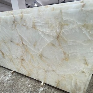 May include: A large slab of white onyx with golden veins. The onyx is polished and smooth, and the veins are intricate and detailed. The onyx is a natural stone and is perfect for use in a variety of applications, including countertops, backsplashes, and flooring.