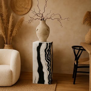 May include: A white and black marble pedestal with a beige ceramic vase with a dried branch arrangement on top. The pedestal is in a room with a beige wall, a woven rug, and a white chair.
