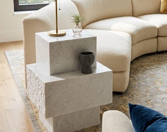 May include: A white marble side table composed of three stacked cubic blocks with varying textures, including smooth and rough-hewn finishes. The table supports a brass lamp with a beige shade, a small glass vase with a plant, and a dark gray ceramic vessel.