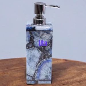 May include: A rectangular soap dispenser with a stainless steel pump. The dispenser is made of a stone material with a blue and white marbled pattern. The dispenser is on a wooden surface.