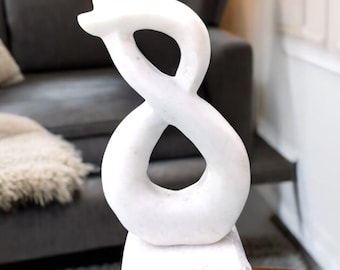 Puede incluir: Escultura de mármol blanco del número ocho, con un diseño abstracto moderno. La escultura está sobre una base de mármol blanco y está sentada sobre una mesa de madera.