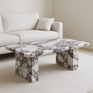 May include: A rectangular coffee table with a marble top featuring a white and purple veined pattern. The table has a solid, block-like base. The table is in front of a white sofa and a white rug.