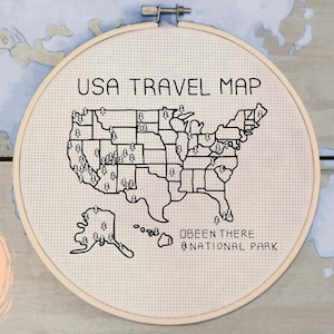 May include: A cross-stitch embroidery hoop featuring a black and white USA travel map. The text "USA TRAVEL MAP" is stitched above the map. The words "Been There National Park" are stitched below the map. The hoop is set against a light wood background.
