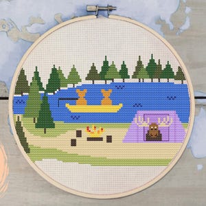 May include: Cross stitch pattern featuring a camping scene with a lake, trees, a tent, a campfire, and two bears in a canoe. The pattern is in a circular hoop.