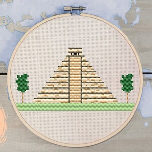 May include: A cross-stitch pattern of a Mayan pyramid with two green trees on either side. The pattern is on a white background and is framed in a wooden hoop. The text "PDF" is written on a peach-colored circle.