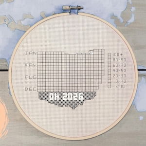 Pode incluir: Um design de ponto cruz em um bastidor de madeira com o contorno de Ohio, com um padrão de grade e o texto "OH 2026". O design inclui uma escala e os meses do ano.