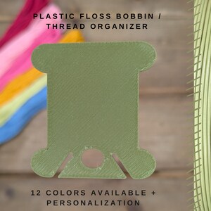 May include: A green plastic floss bobbin thread organiser. The text "PLASTIC FLOSS BOBBIN / THREAD ORGANIZER" is at the top. The text "12 COLORS AVAILABLE + PERSONALIZATION" is at the bottom. Colourful embroidery floss is in the background.