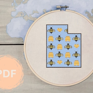 Könnte beinhalten: Ein blauer und weißer Kreuzstich-Stickrahmen mit einem Motiv des Bundesstaates Utah, gefüllt mit gelben und schwarzen Bienen. Der Rahmen befindet sich auf einer Holzoberfläche.
