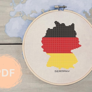 May include: A cross-stitch pattern of the outline of Germany in the colors of the German flag: black, red, and yellow. The pattern is on a white canvas in a wooden embroidery hoop. The text "GERMANY" is printed below the pattern.