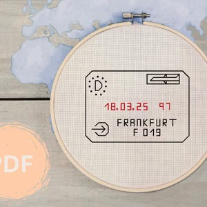 Puede incluir: Un aro de bordado de punto de cruz blanco con un diseño en blanco y negro de una tarjeta de embarque de vuelo. La tarjeta de embarque tiene el texto "18.03.25 97 FRANKFURT F 019" en rojo.