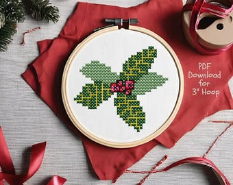 May include: A cross-stitch pattern in a 3-inch wooden embroidery hoop featuring a holly leaf design with green and yellow geometric details and red berries. The white fabric displays the text PDF Download for 3" Hoop in a simple font.