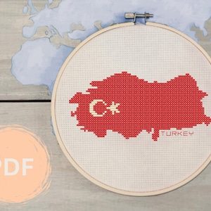 Può includere: Un motivo a punto croce rosso e bianco del contorno della Turchia con una stella e una mezzaluna al centro. Il motivo è su un tessuto bianco e si trova in un cerchio da ricamo in legno. Il testo "TURKEY" è sotto il motivo.