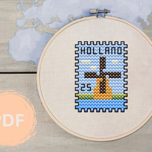 May include: A white embroidery hoop with a cross-stitch design of a windmill with the word "HOLLAND" above it. The design is on a blue background with white clouds.