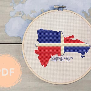 Puede incluir: Un aro de bordado blanco con un diseño de punto de cruz de la bandera de la República Dominicana en forma de país. El texto "REPÚBLICA DOMINICANA" está debajo de la bandera.