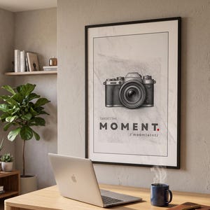 May include: A framed wall art print featuring a black and white illustration of a camera centered above the text "TARGET THE MOMENT." with a phonetic pronunciation guide below, all set against a light gray textured background.