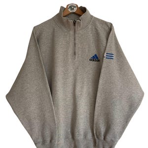 Könnte beinhalten: Ein grauer Half-Zip-Pullover mit dem Adidas-Logo und einem blauen Patch am Ärmel, auf dem "adidas equipment" steht.