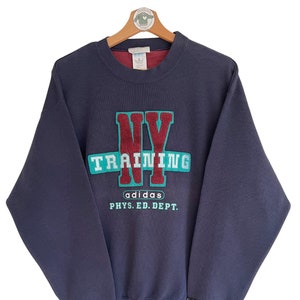 Könnte beinhalten: Ein marineblauer Sweatshirt mit einem gestickten roten, weißen und türkisfarbenen Grafik, auf dem "NY TRAINING adidas PHYS. ED. DEPT." steht.