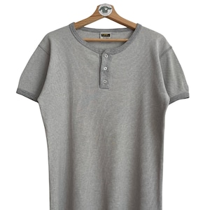 Könnte beinhalten: Hellgraues Henley-Shirt mit kurzen Ärmeln, gerippter Struktur und einer Knopfleiste mit drei Knöpfen. Das Shirt hat einen kontrastierenden grauen Besatz am Kragen und an den Ärmeln. Das Shirt hängt an einem Holzbügel.