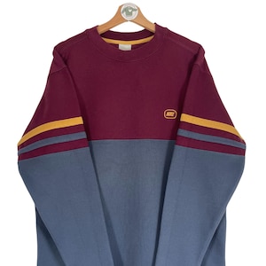 Könnte beinhalten: Ein langärmliger Sweatshirt in den Farben Rotbraun und Blau mit gelben Streifen an den Ärmeln. Der Sweatshirt hat ein kleines "NIKE"-Logo auf der Brust.