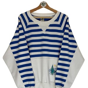 Könnte beinhalten: Ein weiß-blaues gestreiftes Sweatshirt mit einem Segelboot-Motiv auf der Vorderseite. Das Sweatshirt hat einen Rundhalsausschnitt und lange Ärmel. Der Markenname "Puma" ist auf dem Etikett sichtbar.