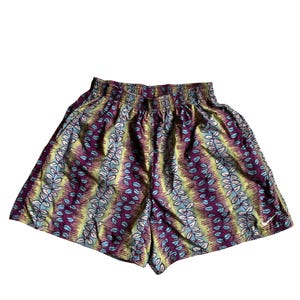 Könnte beinhalten: Sportshorts mit elastischem Bund und einem lebendigen, sich wiederholenden Muster aus vertikalen Streifen in Violett-, Gelb- und Hellblautönen mit blattartigen Motiven. Ein kleines weißes Nike-Swoosh-Logo ist am rechten unteren Bein zu sehen.