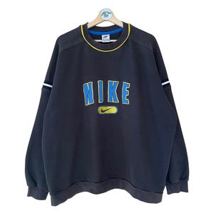 Könnte beinhalten: Dunkelbraunes Nike Sweatshirt mit dem Markennamen in blauen und gelben Buchstaben auf der Brust. Das Sweatshirt hat eine gelb-weiße Einfassung am Kragen und an den Bündchen sowie weiße Streifen an den Ärmeln.