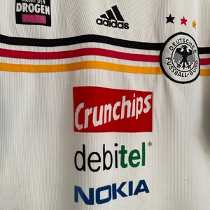 Puede incluir: Camiseta de f&uacute;tbol blanca de Alemania con el logo negro de Adidas, rayas negras y rojas, y el texto "Crunchips", "debitel" y "Nokia". La camiseta tambi&eacute;n presenta el texto "Keine Macht den Drogen" y un escudo de f&uacute;tbol alem&aacute;n.