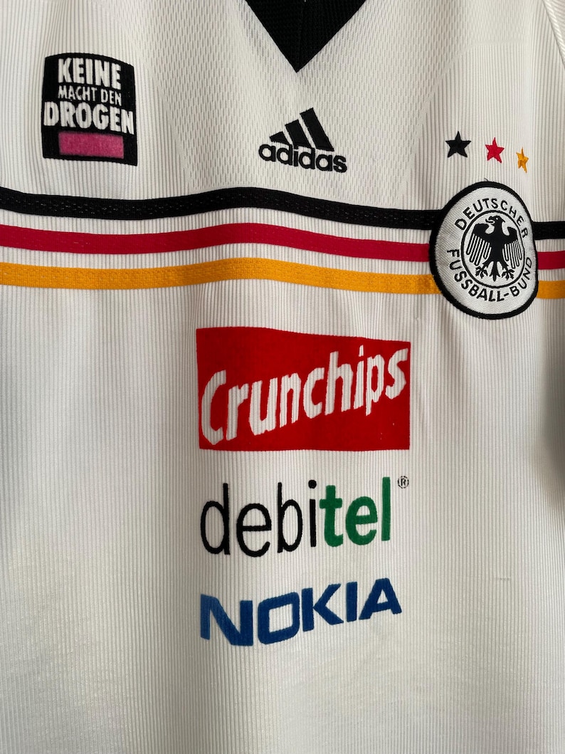 Puede incluir: Camiseta de f&uacute;tbol blanca de Alemania con el logo negro de Adidas, rayas negras y rojas, y el texto "Crunchips", "debitel" y "Nokia". La camiseta tambi&eacute;n presenta el texto "Keine Macht den Drogen" y un escudo de f&uacute;tbol alem&aacute;n.