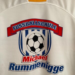 Puede incluir: Camiseta de f&uacute;tbol blanca con el texto "FUSSBALLSCHULE" en una pancarta roja, un gr&aacute;fico de bal&oacute;n de f&uacute;tbol y el nombre "Michael Rummenigge" en rojo y azul. La camiseta tiene una textura acanalada y un cuello con rayas negras, rojas, amarillas y negras.