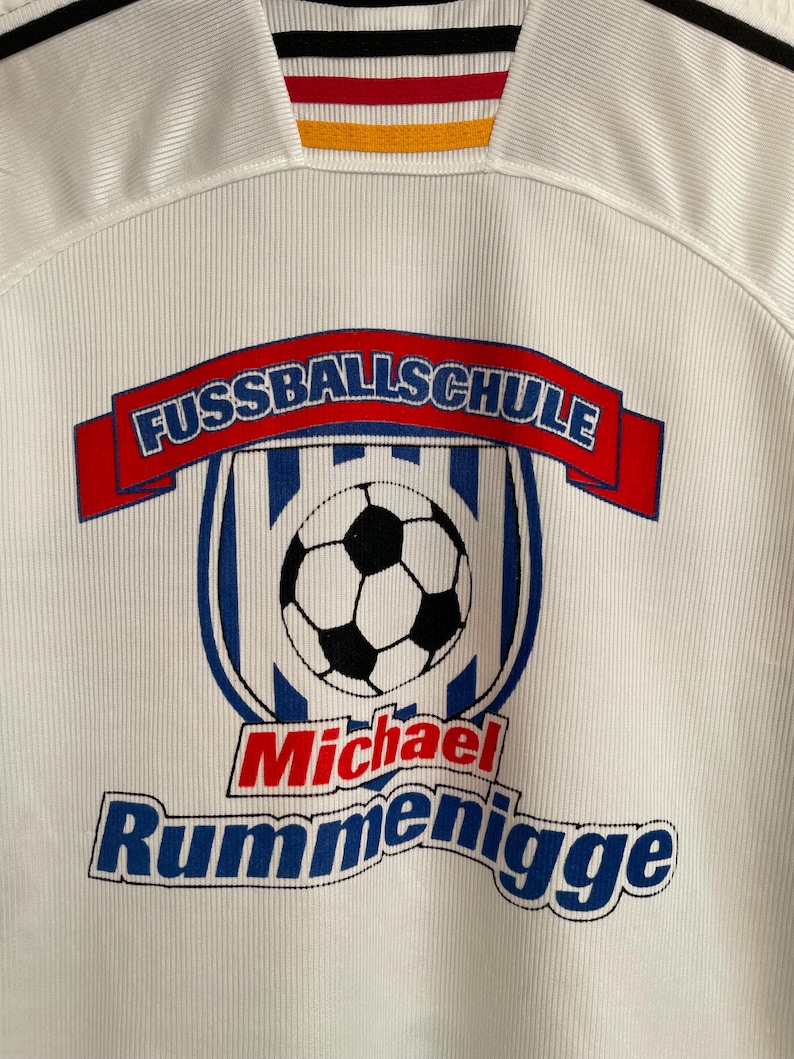 Puede incluir: Camiseta de f&uacute;tbol blanca con el texto "FUSSBALLSCHULE" en una pancarta roja, un gr&aacute;fico de bal&oacute;n de f&uacute;tbol y el nombre "Michael Rummenigge" en rojo y azul. La camiseta tiene una textura acanalada y un cuello con rayas negras, rojas, amarillas y negras.