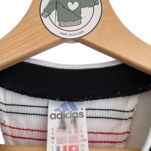 Puede incluir: Una camiseta Adidas vintage con cuello negro y cuerpo blanco, acentuada con rayas rojas y amarillas. La etiqueta Adidas muestra tallas y banderas internacionales. La camiseta est&aacute; colgada en una percha de madera con una pegatina 'Home Alone Vintage'.