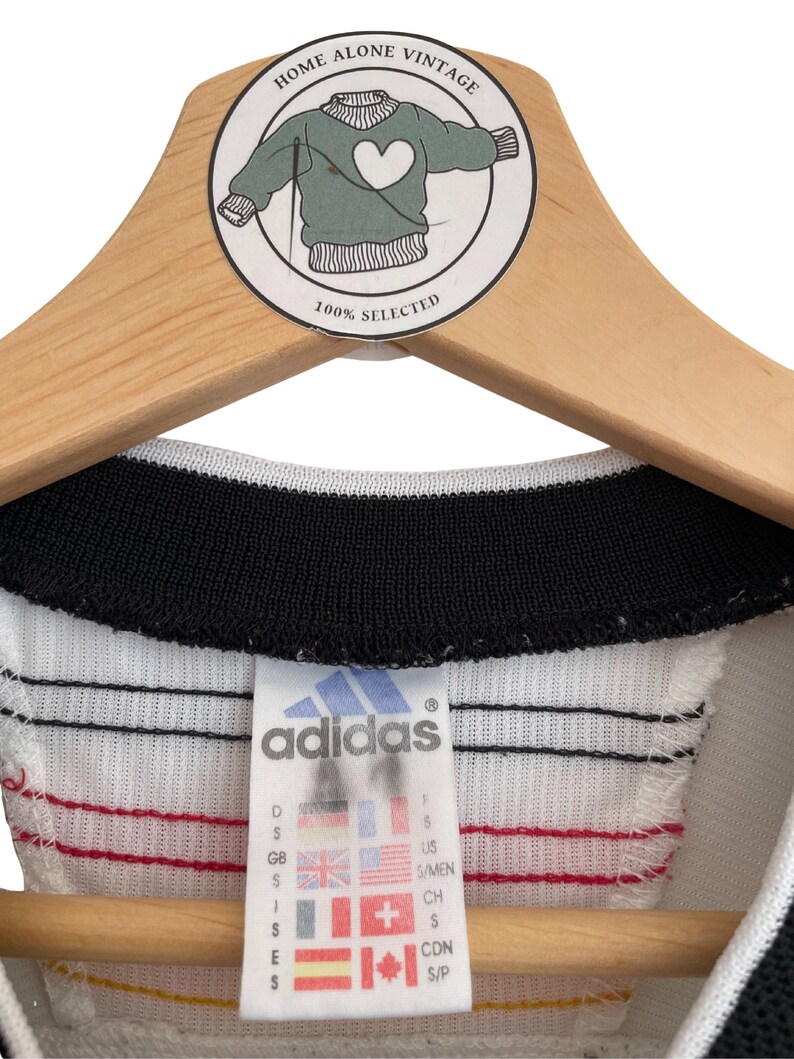 Puede incluir: Una camiseta Adidas vintage con cuello negro y cuerpo blanco, acentuada con rayas rojas y amarillas. La etiqueta Adidas muestra tallas y banderas internacionales. La camiseta est&aacute; colgada en una percha de madera con una pegatina 'Home Alone Vintage'.