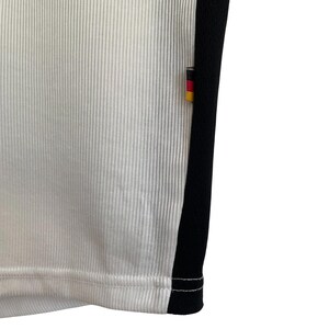 Puede incluir: Primer plano de una camiseta deportiva blanca con textura acanalada vertical. Una franja negra recorre el lateral, con un peque&ntilde;o emblema de la bandera alemana. El dobladillo de la camiseta es visible, mostrando las costuras.