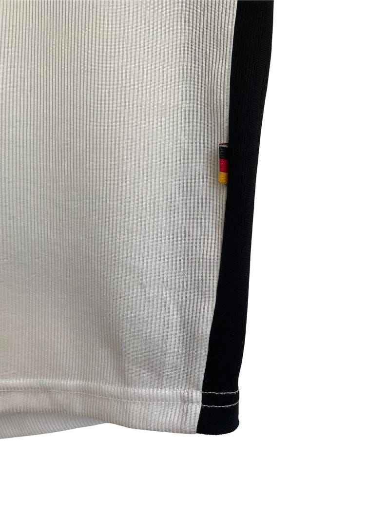 Puede incluir: Primer plano de una camiseta deportiva blanca con textura acanalada vertical. Una franja negra recorre el lateral, con un peque&ntilde;o emblema de la bandera alemana. El dobladillo de la camiseta es visible, mostrando las costuras.