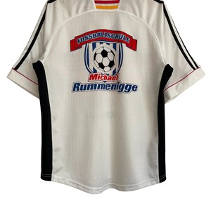 Puede incluir: Camiseta de f&uacute;tbol blanca con detalles negros y rojos. La camiseta presenta el texto "FUSSBALLSCHULE" sobre un gr&aacute;fico de bal&oacute;n de f&uacute;tbol y el nombre "Michael Rummenigge" en azul.