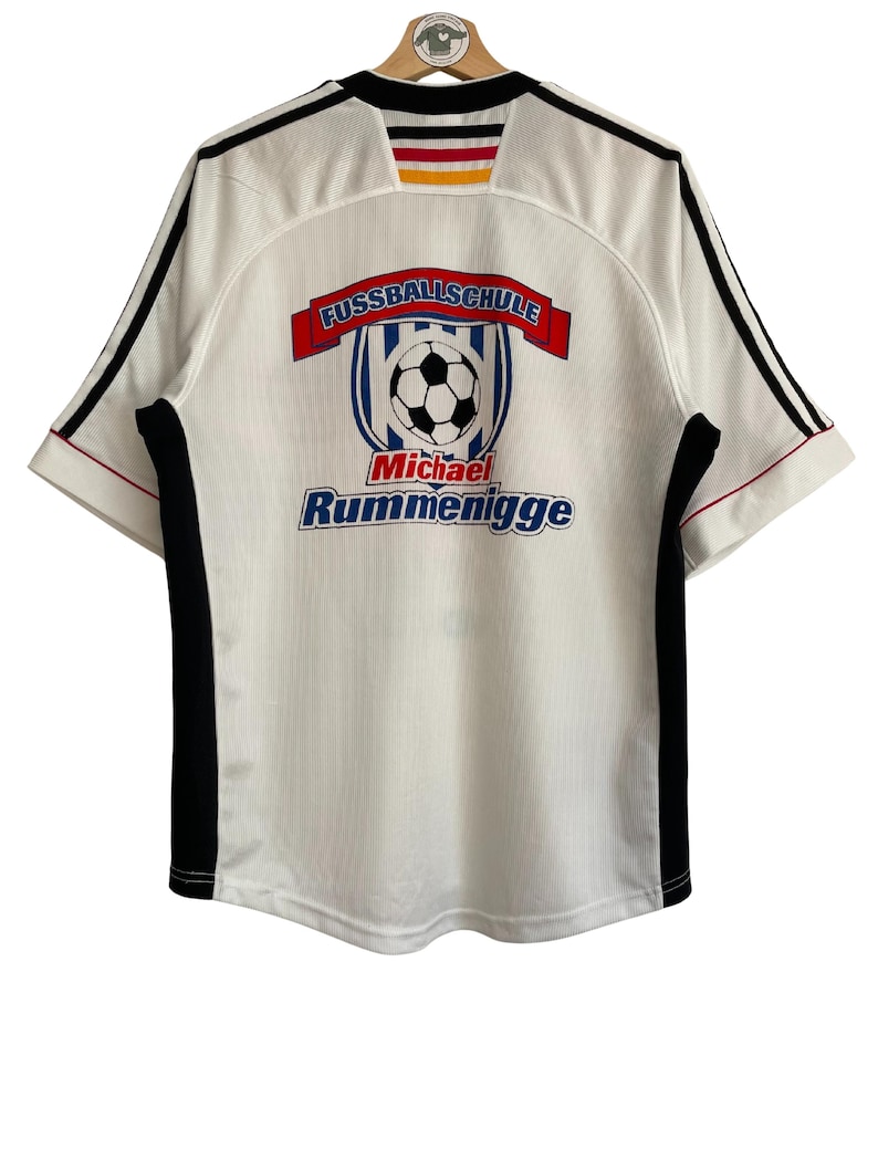 Puede incluir: Camiseta de f&uacute;tbol blanca con detalles negros y rojos. La camiseta presenta el texto "FUSSBALLSCHULE" sobre un gr&aacute;fico de bal&oacute;n de f&uacute;tbol y el nombre "Michael Rummenigge" en azul.