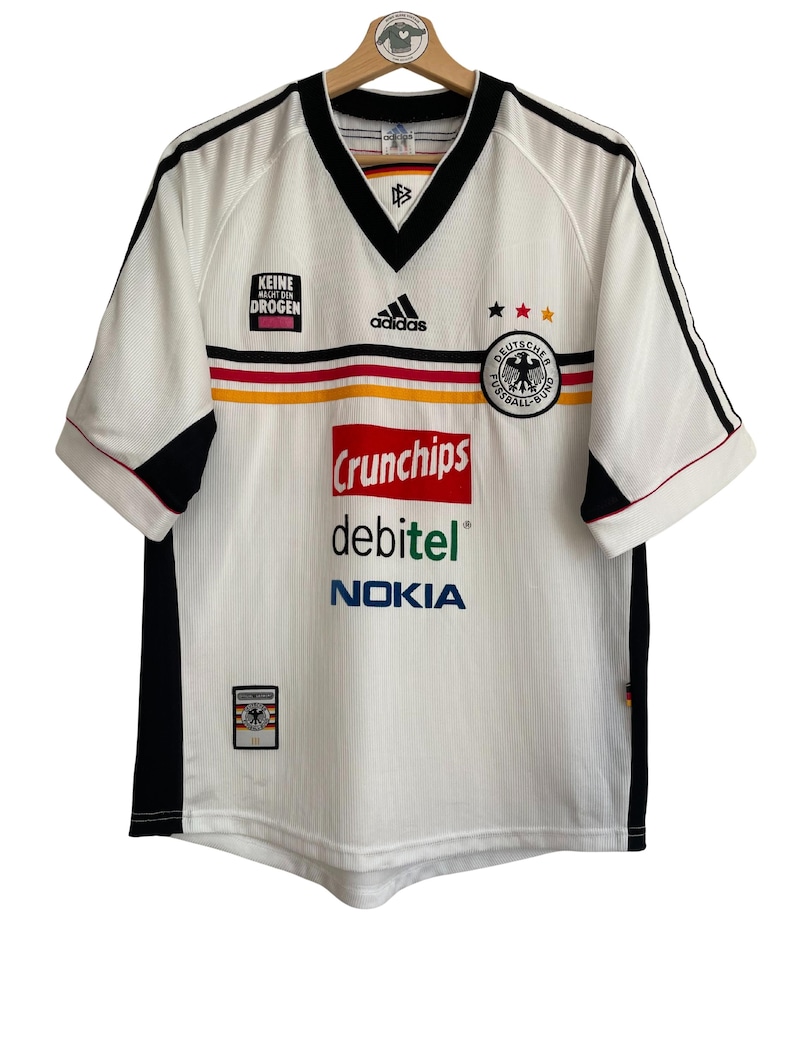 Puede incluir: Camiseta de f&uacute;tbol blanca con detalles negros, cuello en V y el logo de Adidas. Presenta los colores de la bandera alemana y los textos "KEINE MACHT DEN DROGEN", "Crunchips", "debitel" y "NOKIA".
