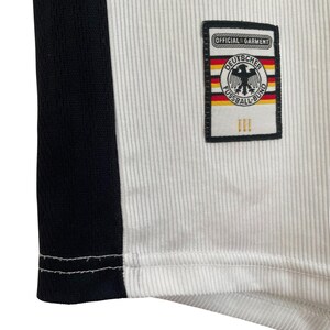 Puede incluir: Primer plano de una camiseta de f&uacute;tbol blanca y negra. La camiseta presenta una franja negra en el lateral y un parche con las palabras "OFFICIAL GARMENT" y "DEUTSCHER FUSSBALL-BUND" con un emblema de &aacute;guila alemana.