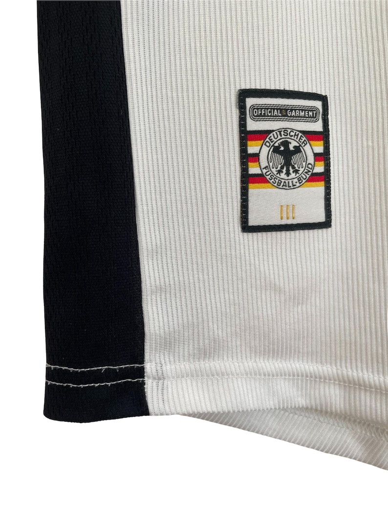 Puede incluir: Primer plano de una camiseta de f&uacute;tbol blanca y negra. La camiseta presenta una franja negra en el lateral y un parche con las palabras "OFFICIAL GARMENT" y "DEUTSCHER FUSSBALL-BUND" con un emblema de &aacute;guila alemana.