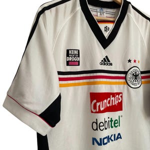 Puede incluir: Camiseta de f&uacute;tbol blanca con detalles negros, con el logo de Adidas y el escudo de la selecci&oacute;n alemana. La camiseta presenta el texto "KEINE MACHT DEN DROGEN" y los logotipos de los patrocinadores Crunchips, debitel y Nokia.