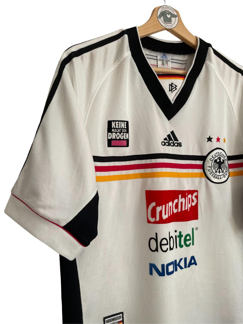 Puede incluir: Camiseta de f&uacute;tbol blanca con detalles negros, con el logo de Adidas y el escudo de la selecci&oacute;n alemana. La camiseta presenta el texto "KEINE MACHT DEN DROGEN" y los logotipos de los patrocinadores Crunchips, debitel y Nokia.