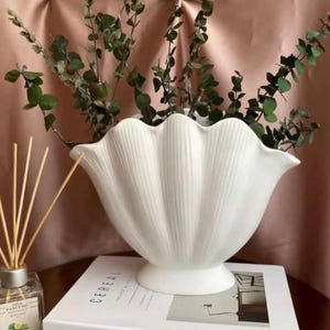May include: White ceramic vase with a scalloped rim and a fluted design. The vase is filled with dried eucalyptus branches and is sitting on a book with the word "CEREAL" printed on the cover.