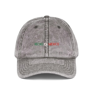 Könnte beinhalten: Eine anthrazitfarbene Baseballkappe mit verwaschener Acid-Wash-Optik. Auf der Vorderseite befindet sich der gestickte Schriftzug HECHO EN MEXICO, wobei HECHO in Grün, EN in Weiß und MEXICO in Rot gehalten ist, passend zur mexikanischen Flagge.