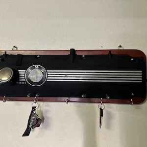 Könnte beinhalten: Ein schwarzer BMW-Motorventildeckel, der als Schlüsselhalter wiederverwendet wurde. Der Schlüsselhalter ist auf einer Holzplatte mit Metallhaken für Schlüssel montiert. Das BMW-Logo ist sichtbar, zusammen mit dem Text "ZUNDFOLGE" und "153624".