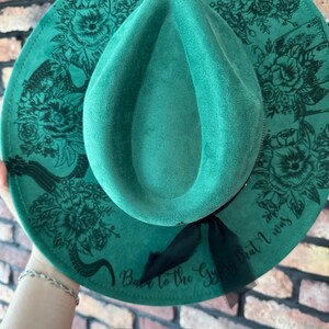Puede incluir: Un sombrero fedora de fieltro verde con una cinta negra y un dise&ntilde;o floral. El sombrero tiene el texto "Back to the Gypsy that I was" impreso en el ala.