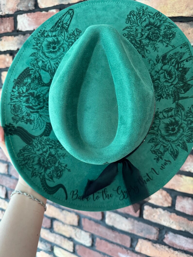 Puede incluir: Un sombrero fedora de fieltro verde con una cinta negra y un dise&ntilde;o floral. El sombrero tiene el texto "Back to the Gypsy that I was" impreso en el ala.
