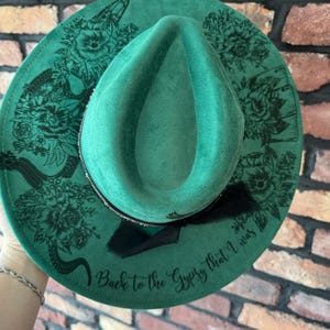 Puede incluir: Un sombrero fedora de fieltro verde con un dise&ntilde;o floral negro y el texto "Back to the Gypsy that I was" en el borde.
