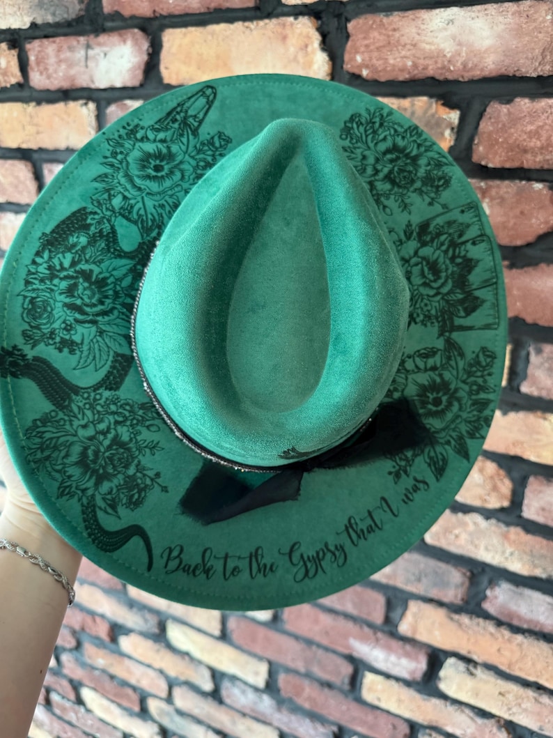 Puede incluir: Un sombrero fedora de fieltro verde con un dise&ntilde;o floral negro y el texto "Back to the Gypsy that I was" en el borde.