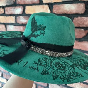 Puede incluir: Un sombrero fedora de fieltro verde con una cinta negra y un dise&ntilde;o de p&aacute;jaro y flores. El sombrero tiene una cinta negra con pedrer&iacute;a y el texto "But I was" impreso en el ala.