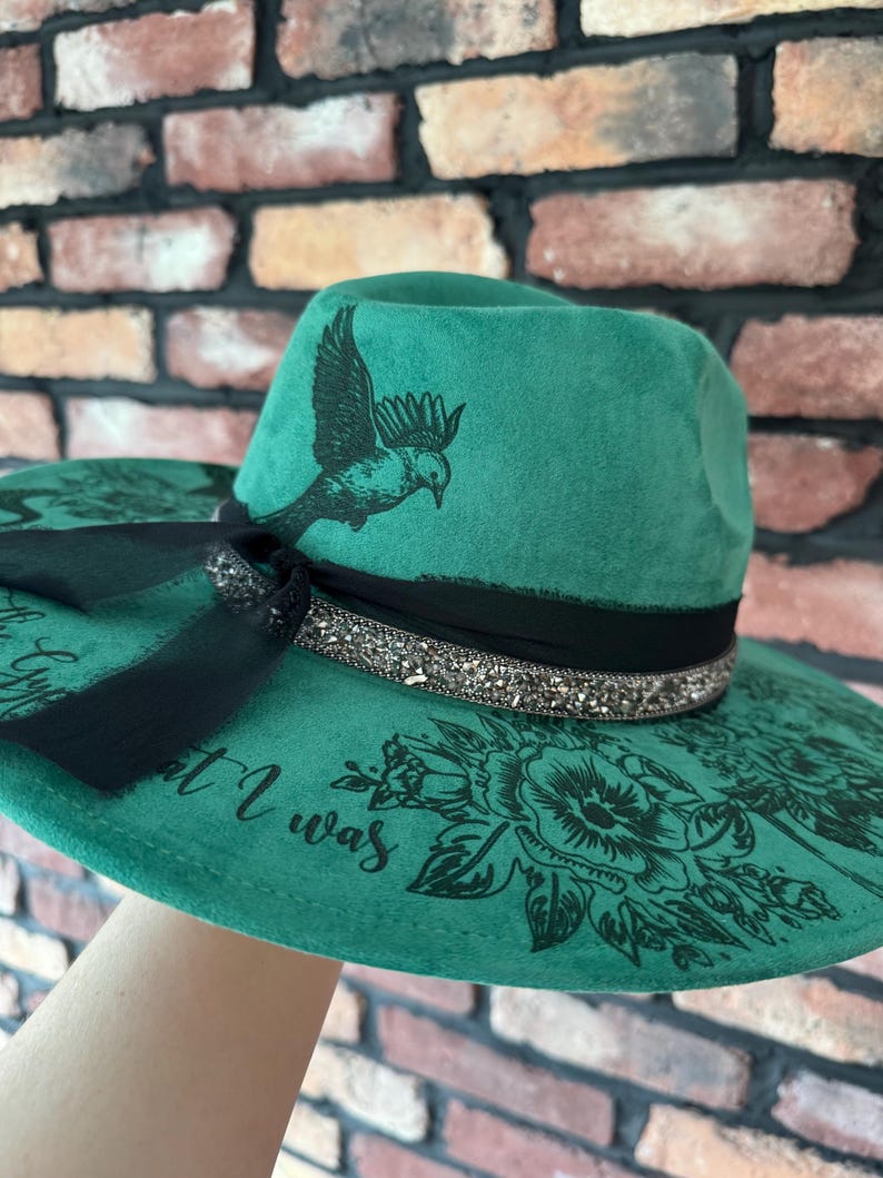 Puede incluir: Un sombrero fedora de fieltro verde con una cinta negra y un dise&ntilde;o de p&aacute;jaro y flores. El sombrero tiene una cinta negra con pedrer&iacute;a y el texto "But I was" impreso en el ala.